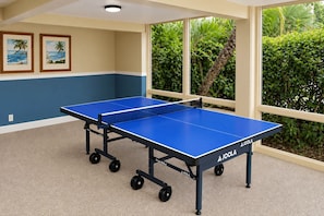 Una habitación con dos mesas de ping pong azules, un ventanal con vistas a zonas verdes y dos cuadros en la pared.