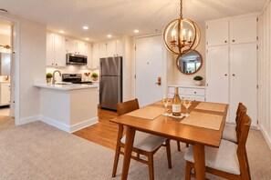 A modern kitchen with white cabinetry, stainless steel appliances, and a dining area with a wooden table and chairs. - Whitewater Ocean View Condo | North Coast Village (Oceanside)