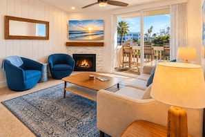 Una sala de estar con chimenea, dos sillones azules, un sofá color beige, una mesa de centro y vista a una playa a través de un ventanal corredero.