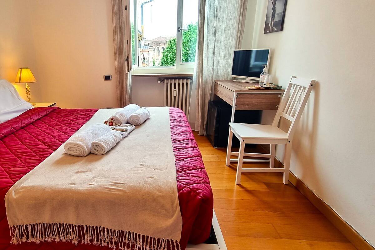 Hotelkamer met een bed, een bureau met een computer, een stoel en een raam met uitzicht op gebouwen.