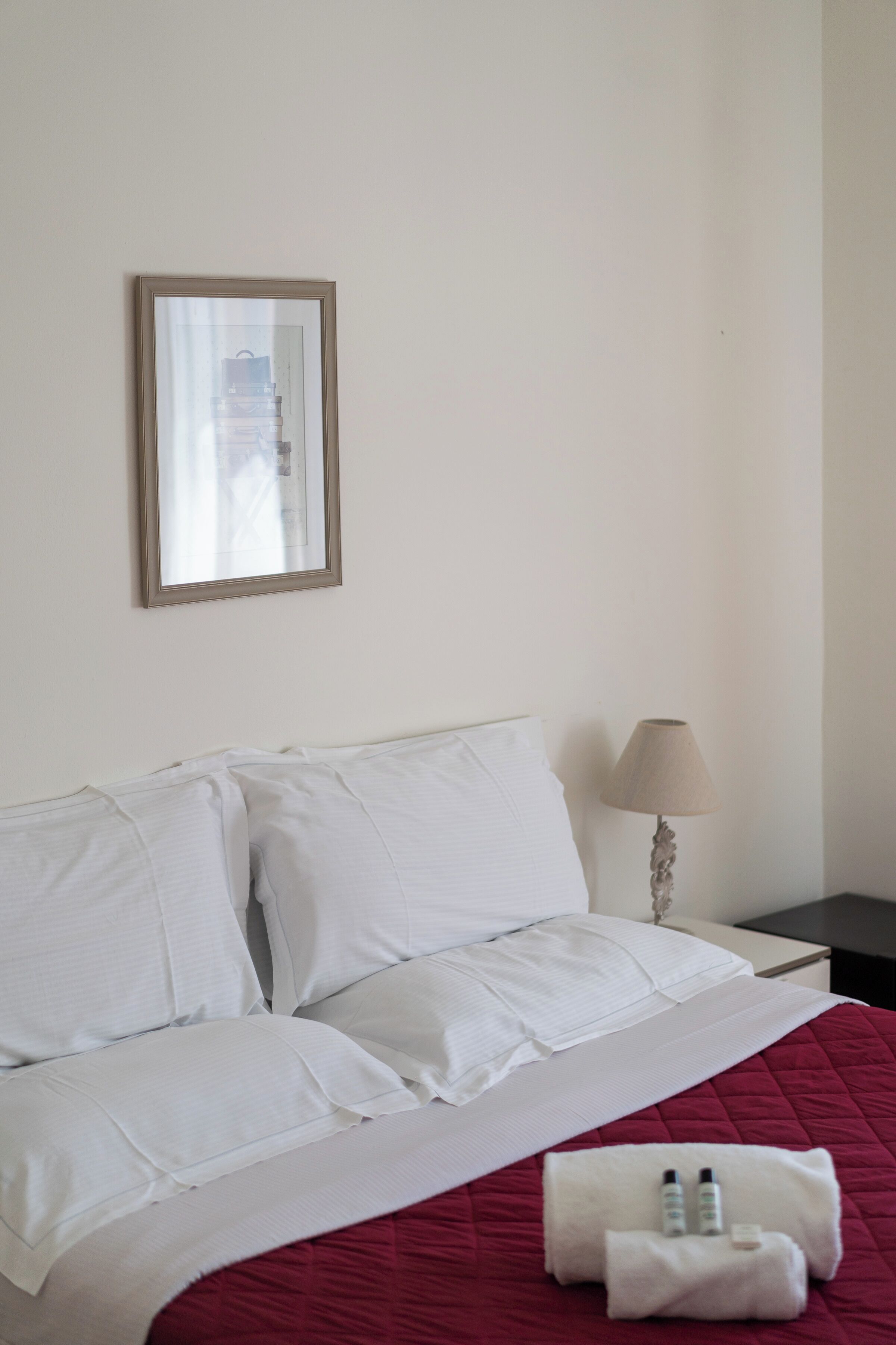 A neatly made bed with white linens and a red quilt, a bedside lamp, and a framed picture on the wall.