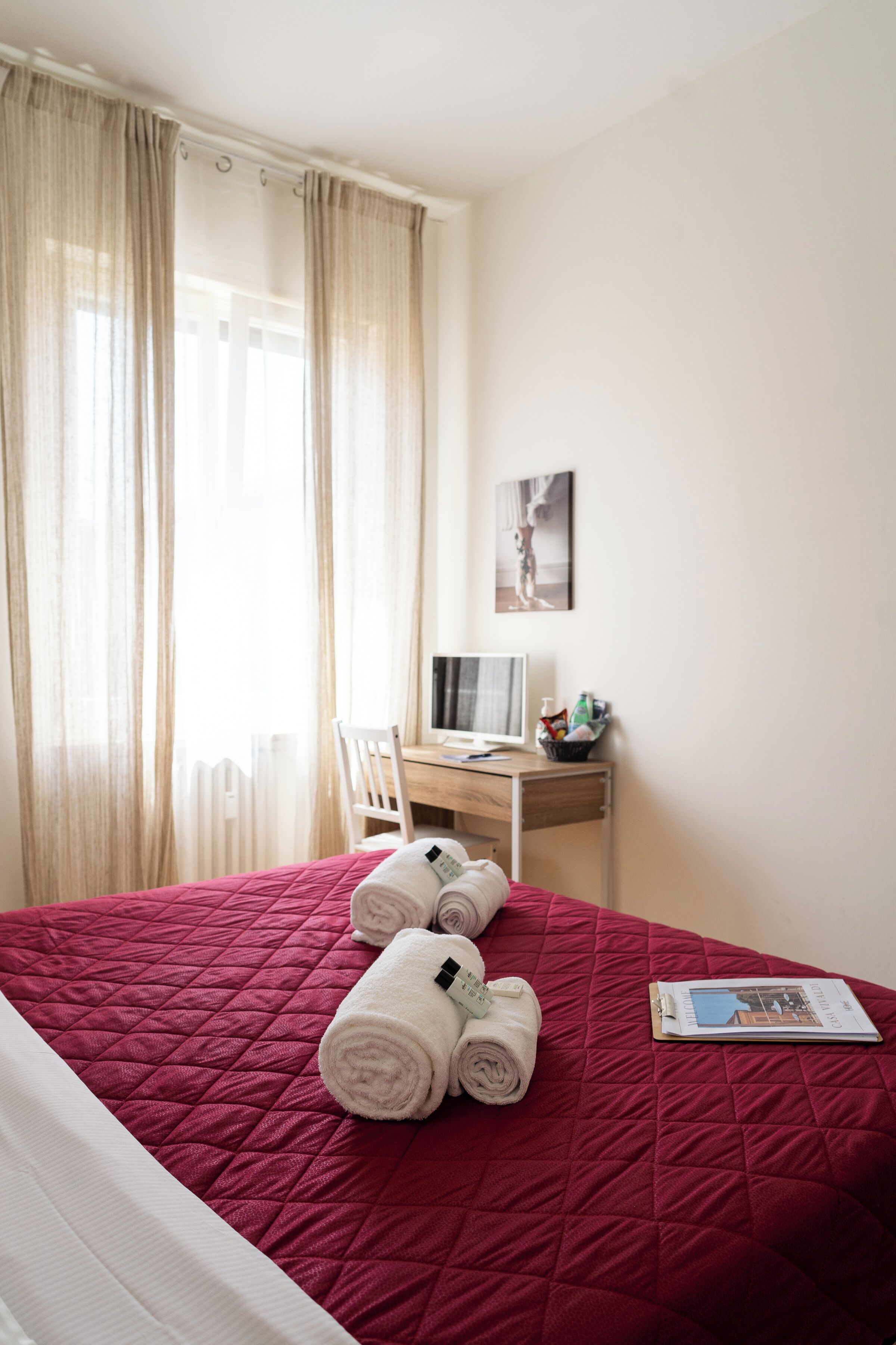 Une chambre d’hôtel avec un couvre-lit rouge matelassé, un bureau avec un ordinateur et une chaise. On trouve des serviettes pliées sur le lit et un tableau accroché au mur.