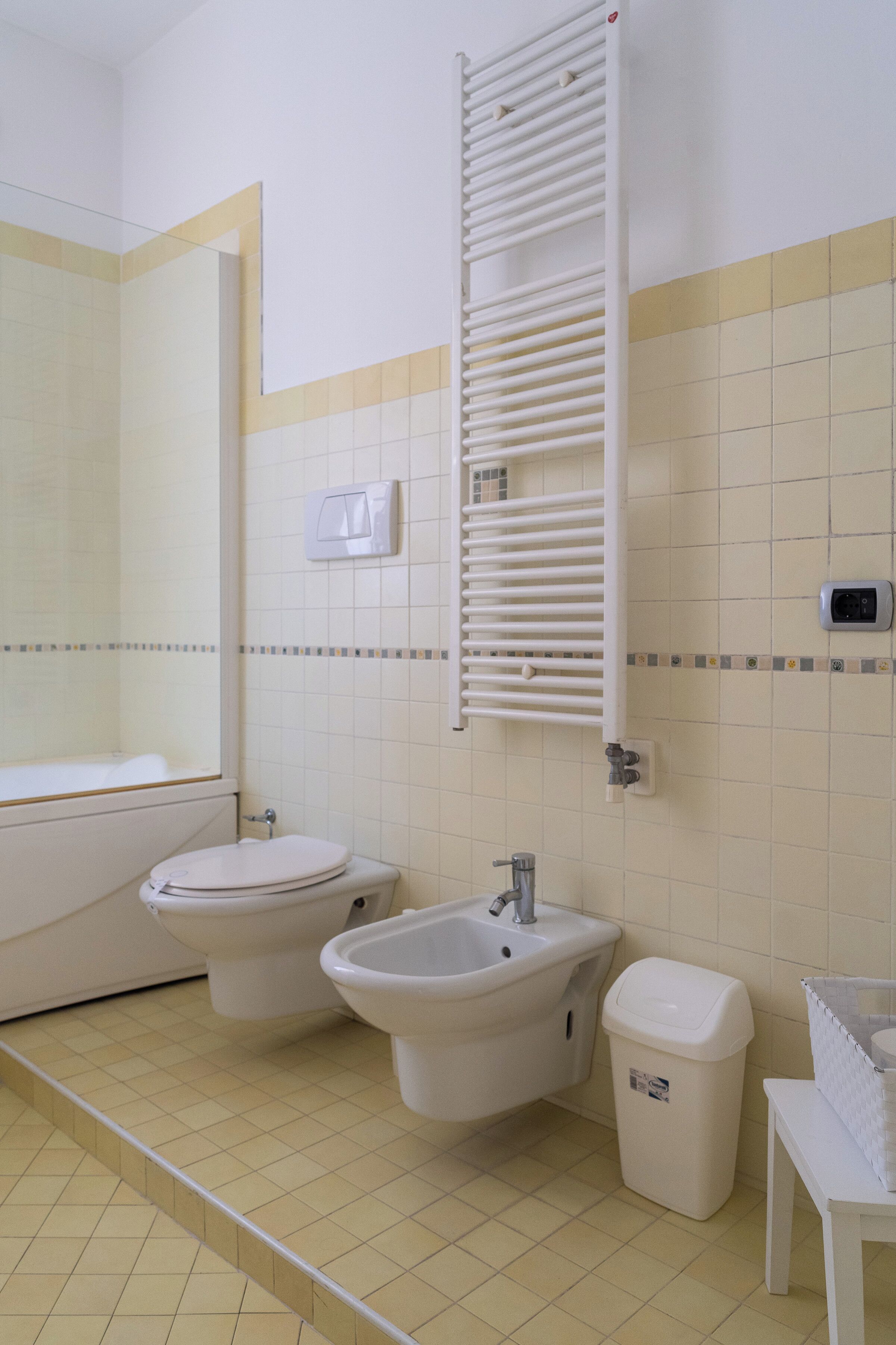A bathroom with two toilets, a sink, a bidet, a towel rack, and a trash bin.