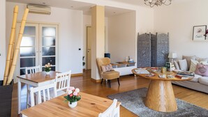 Een moderne woonkamer met een houten eettafel, witte stoelen, een ronde salontafel en een bank. Er is een kroonluchter, een wandairconditioner en een decoratief scherm met ingewikkelde patronen.