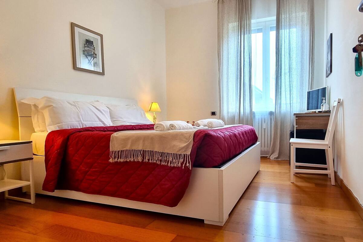Een netjes opgemaakt bed met een rode deken, een nachtkastje met een lamp, een klein bureau met een computer en een raam met dunne gordijnen.
