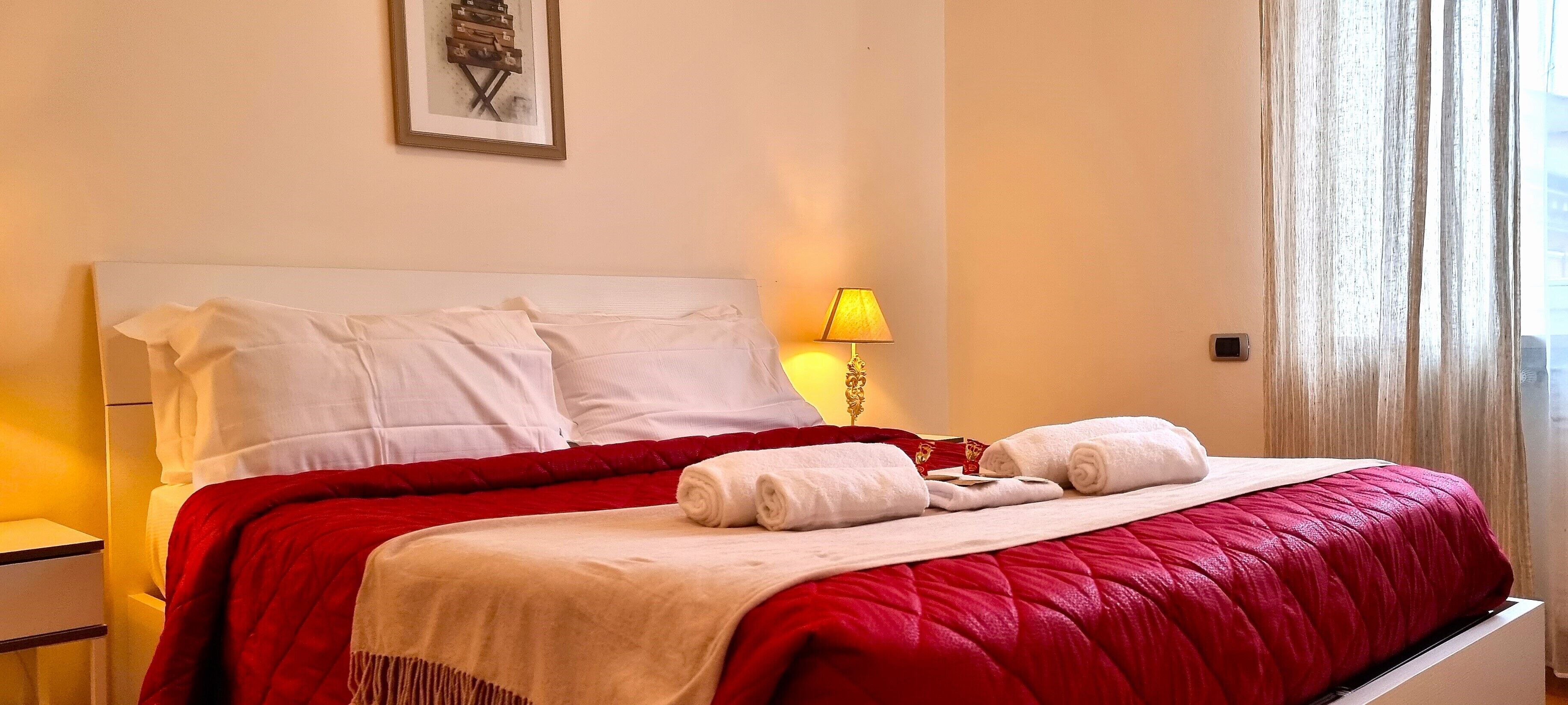 A neatly made bed with white linens and a red quilt, a bedside lamp, and a framed picture on the wall.