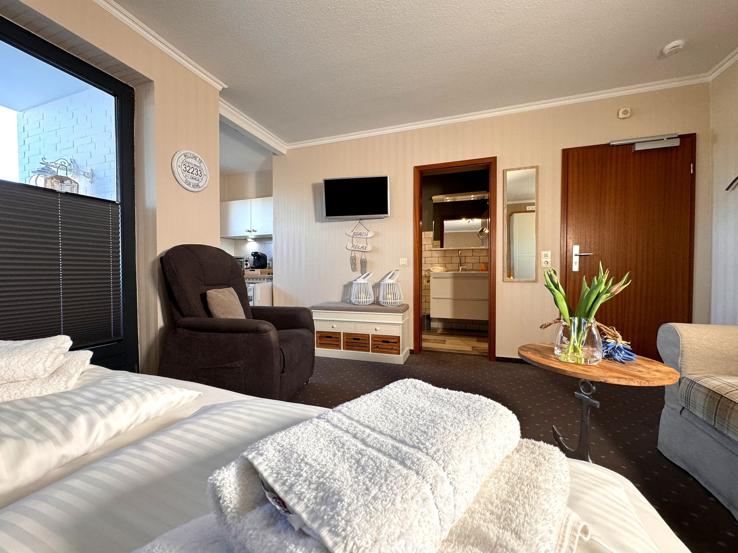 A hotel room with a bed, a chair, a desk, a TV, a bathroom, and a small table with a vase.