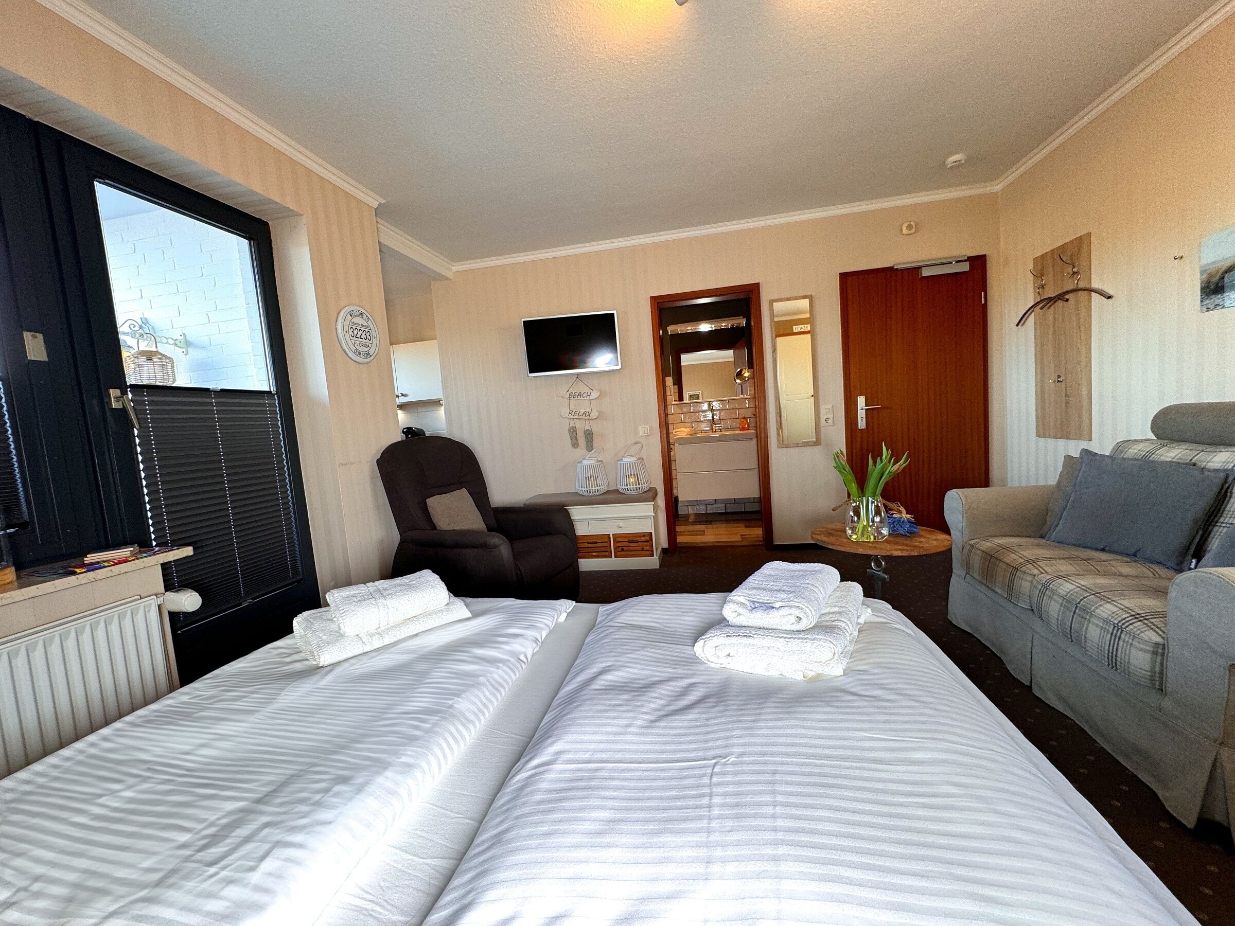 A hotel room with a bed, a chair, a sofa, a TV, and a bathroom visible through an open door.