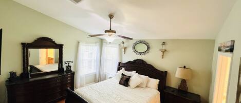 A bedroom with a large bed, a ceiling fan, a dresser, a mirror, and a window with curtains.