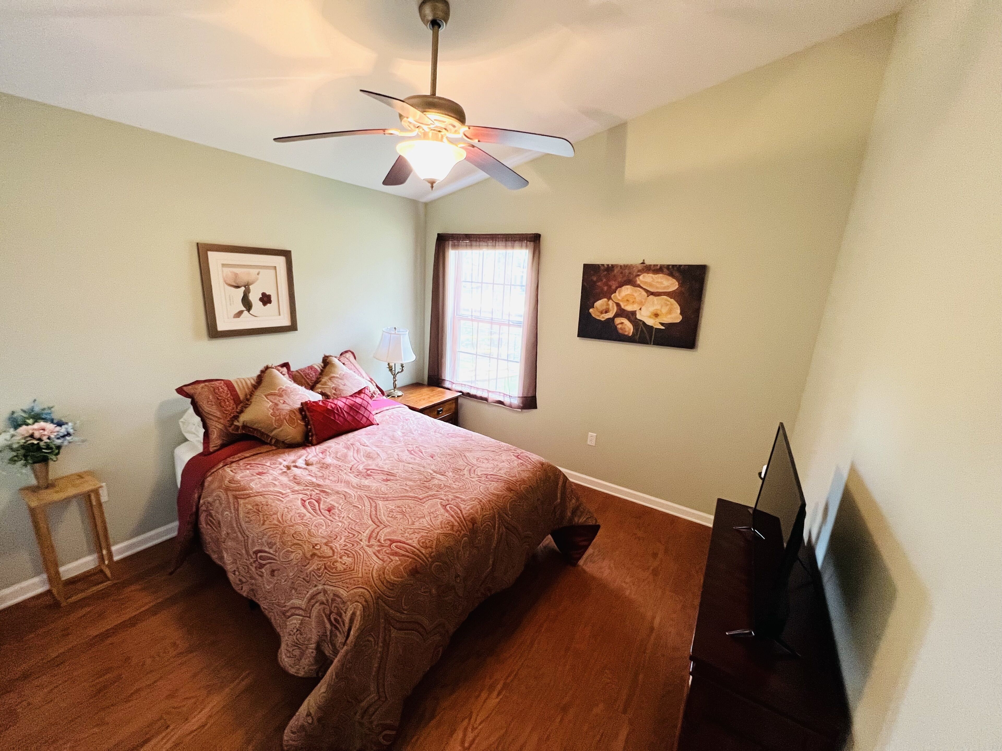 A bedroom with a bed, a ceiling fan, a window, a TV, and artwork on the walls.