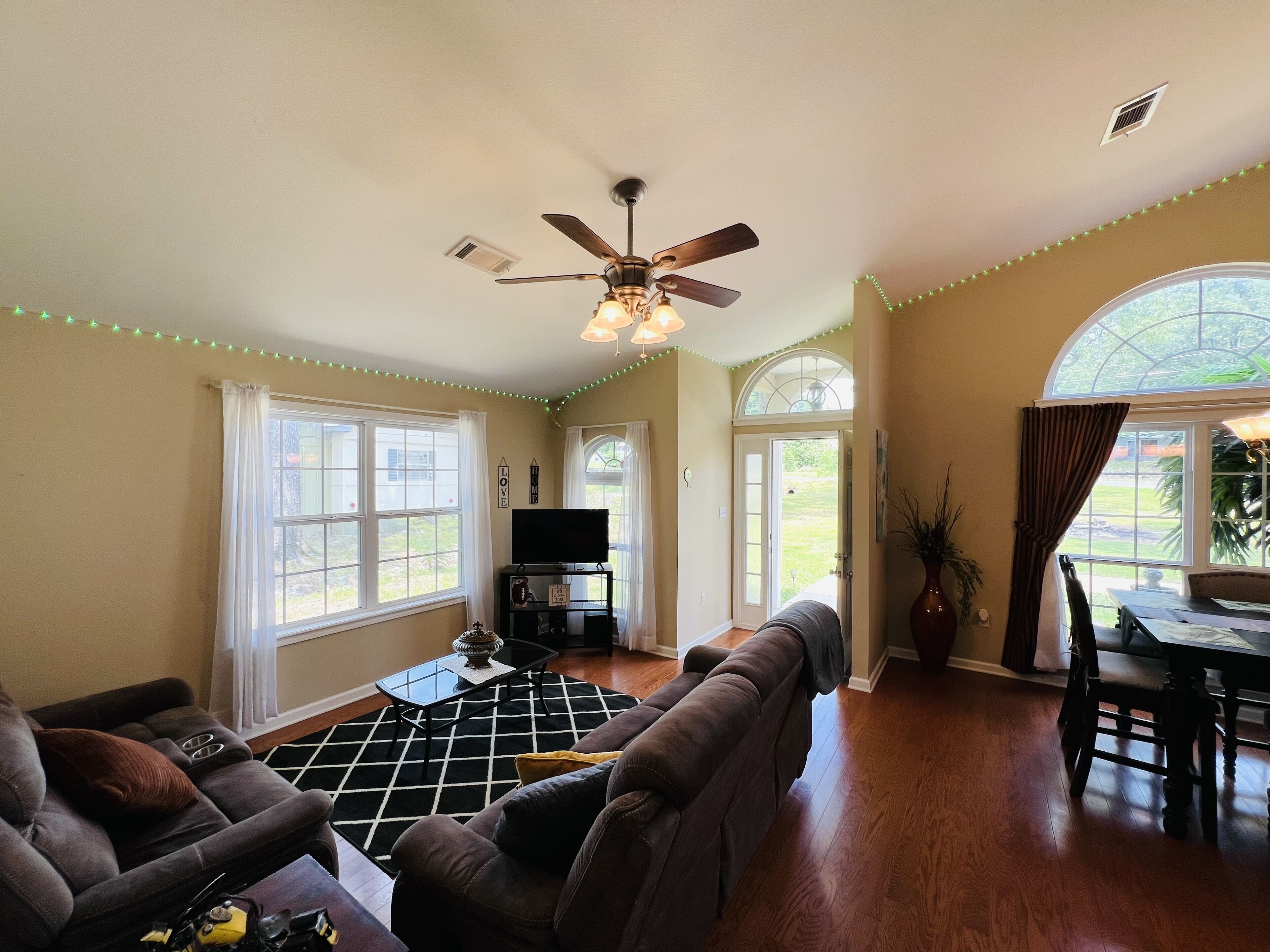 A living room with a ceiling fan, a TV on a stand, a sofa, and a dining area with a table and chairs.