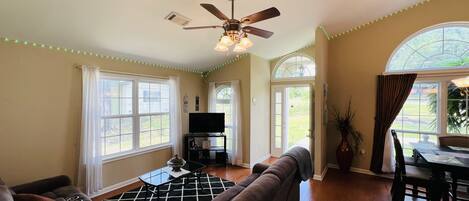 A living room with a ceiling fan, a TV on a stand, a sofa, and a dining area with a table and chairs.