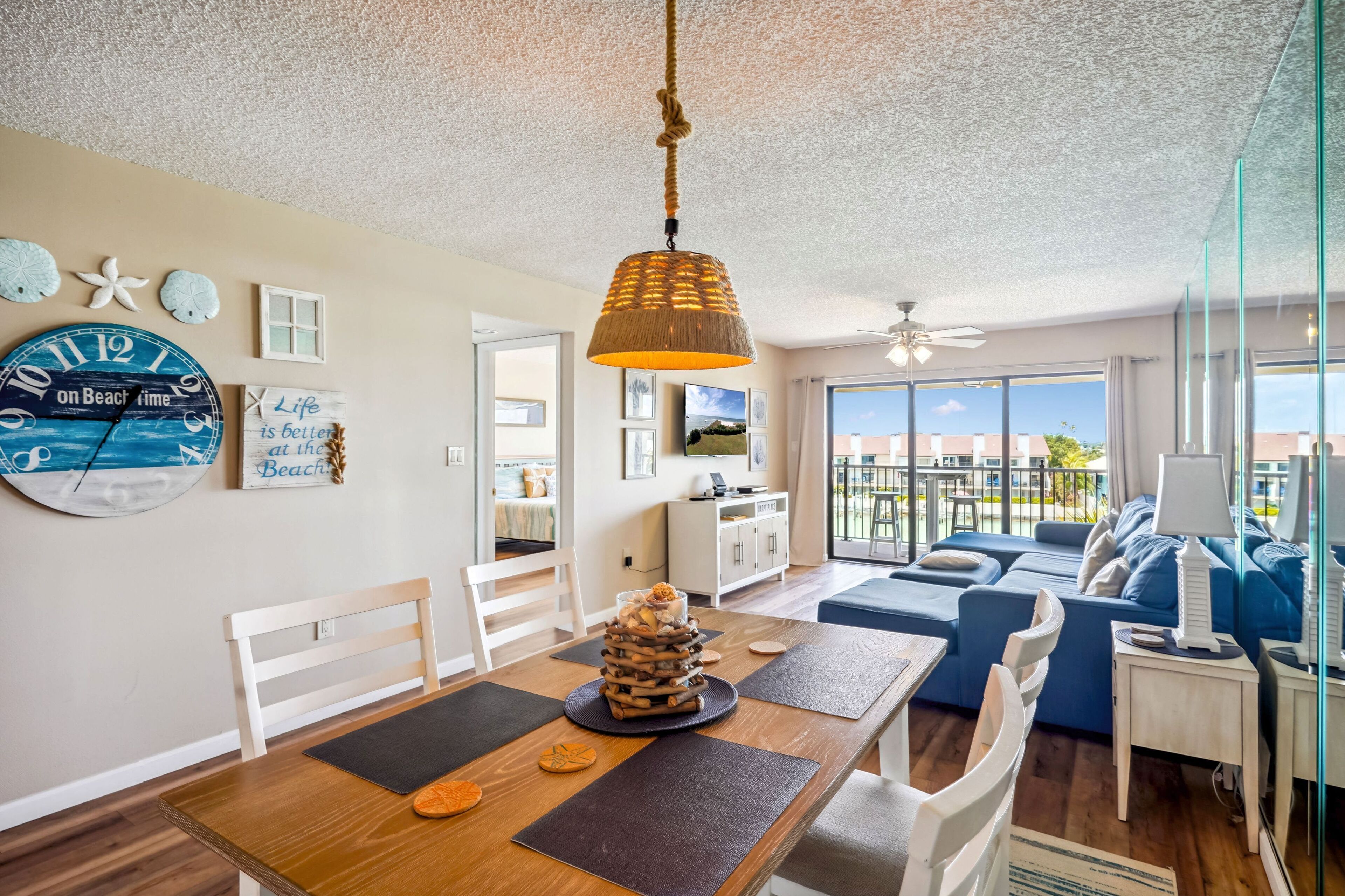 A spacious living area with a dining table, chairs, a hanging light fixture, and a large clock on the wall.