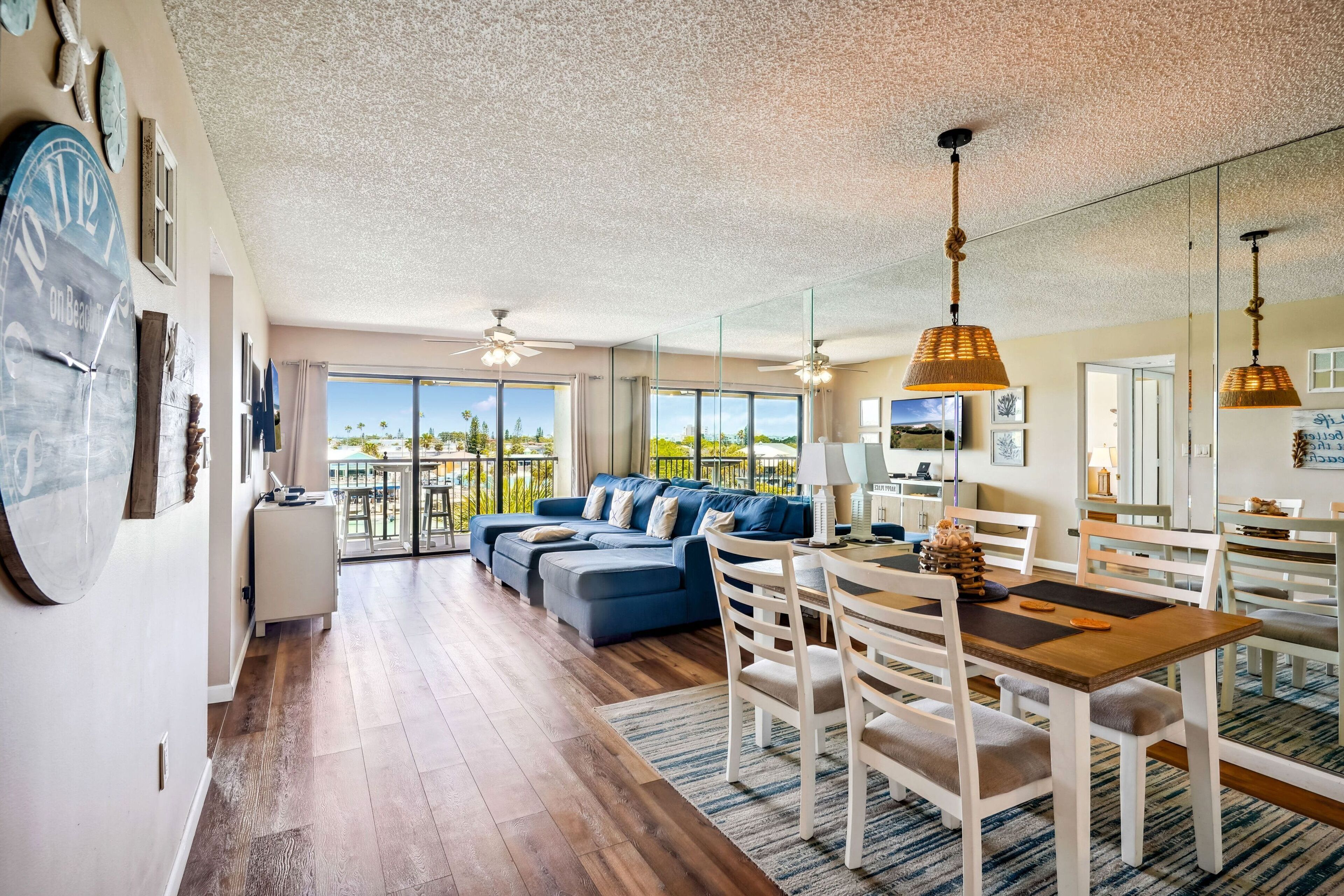A spacious living area with a large sofa, dining table, and chairs. There is a balcony with a view of the outdoors.