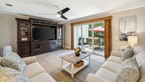 Una sala de estar con un televisor de pantalla plana, un mueble de entretenimiento de madera oscura, sofás color beige, una mesa de centro y un puerta corrediza de vidrio que da a un patio exterior.