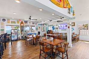 A beach-themed restaurant interior with wooden tables and chairs, a refrigerator with beverages, and a view of the ocean through the windows.