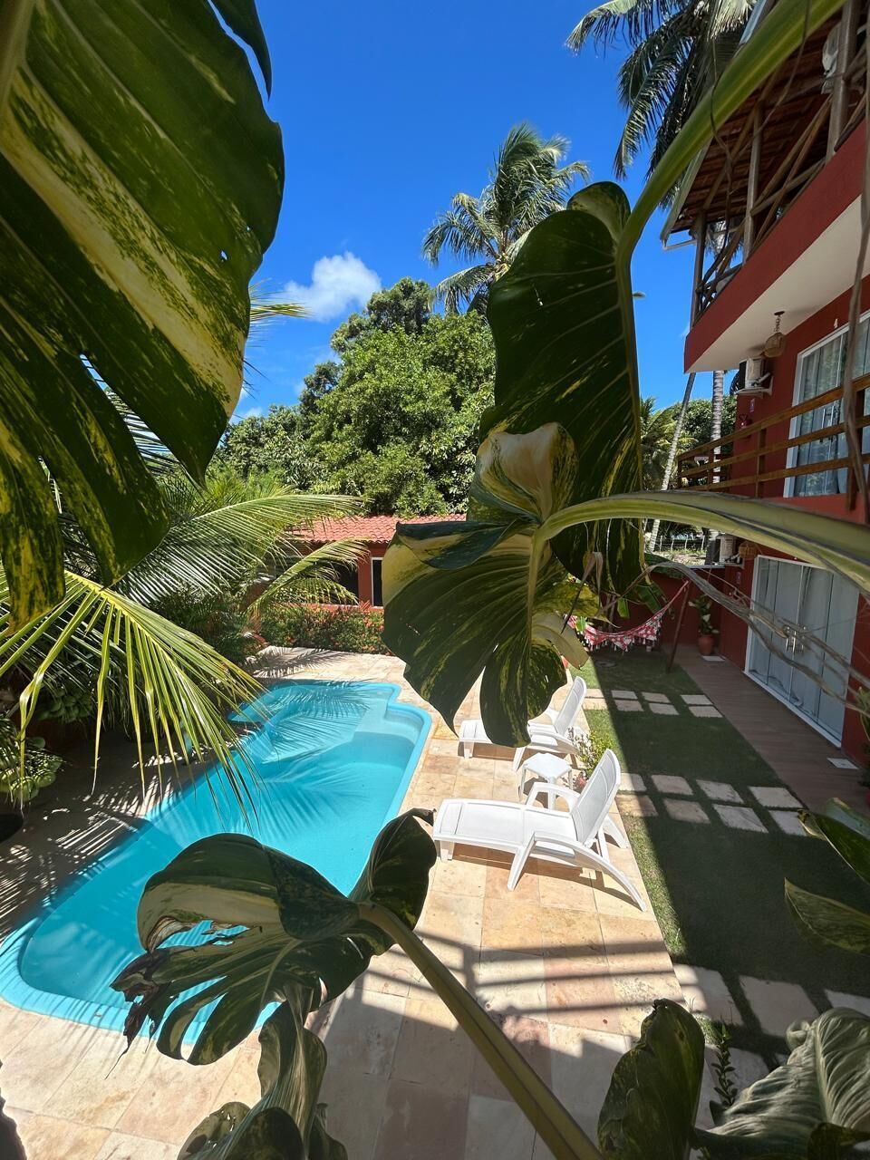 Un espace piscine avec un bâtiment rouge, des chaises blanches et de grandes plantes tropicales.