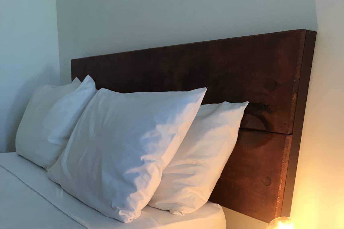 Una cama con ropa de cama y almohadas blancas, un cabecero de madera y una mesita de noche con una lámpara encendida.
