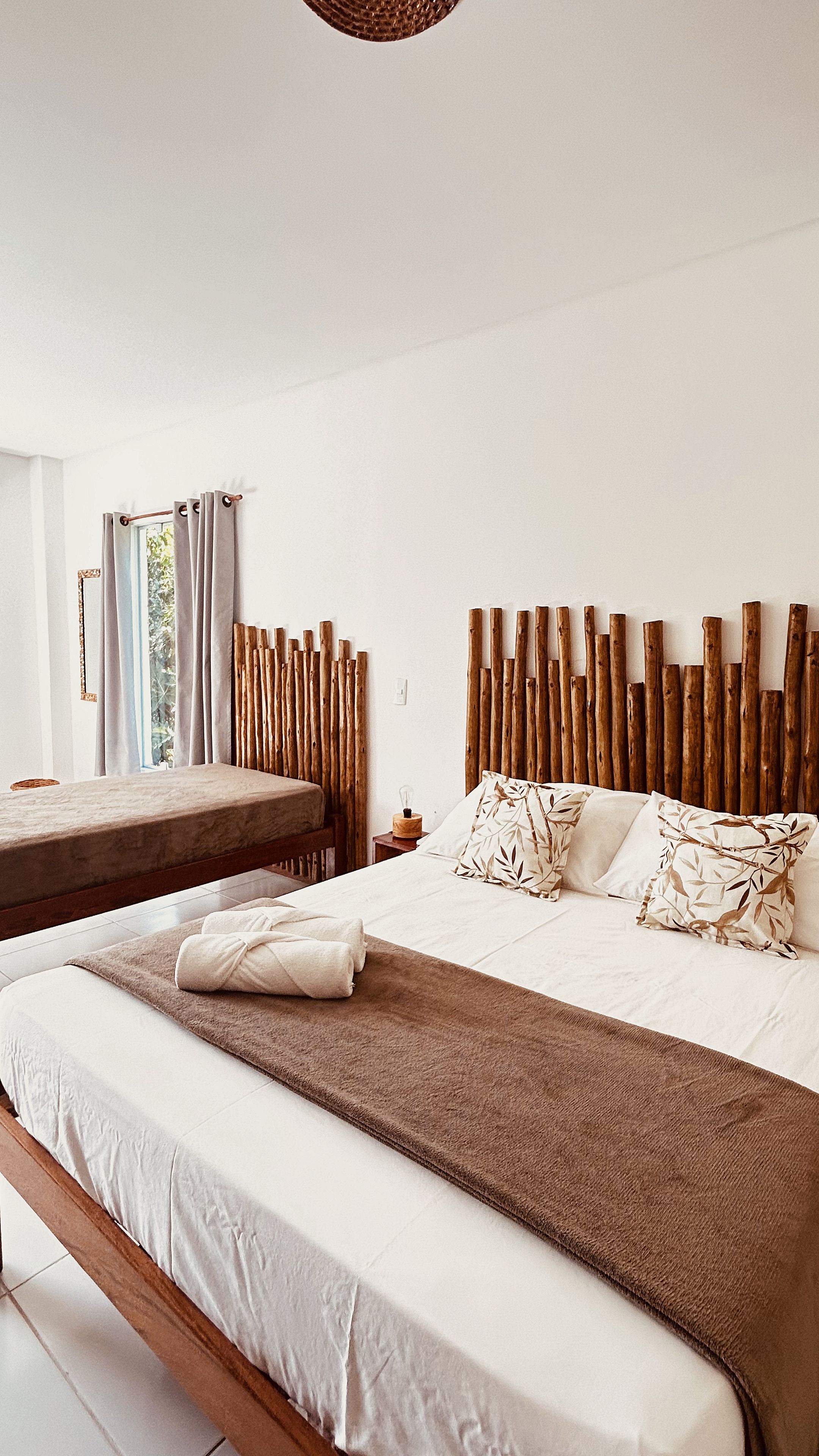 Quarto com uma cama grande, uma cama menor, cabeceira de madeira, mesa de cabeceira e janela com cortinas.