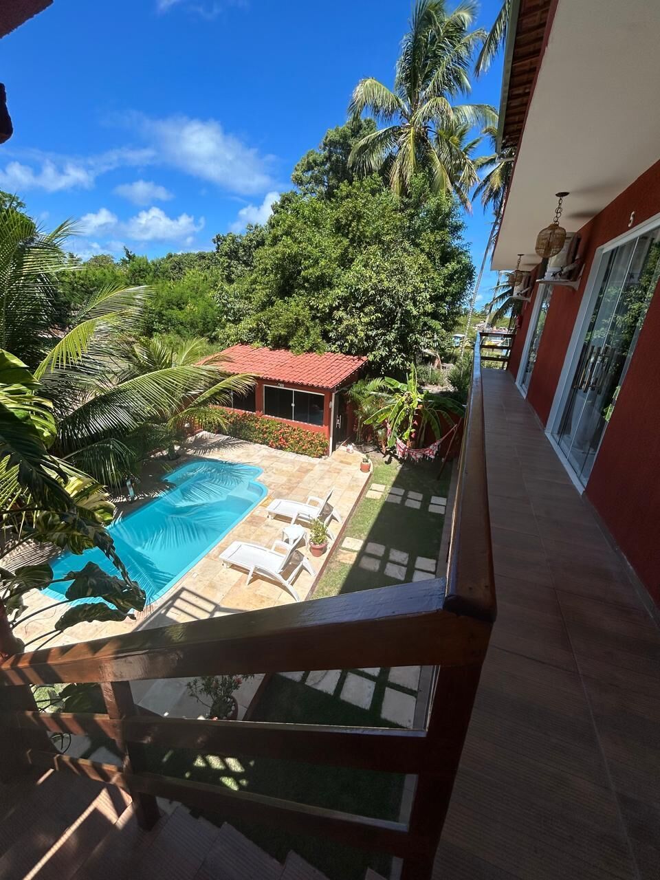 Uma varanda com vista para uma piscina, uma construção com telhado vermelho e vegetação exuberante.