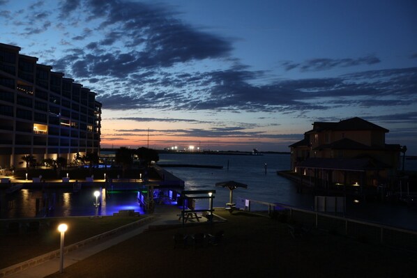 A waterfront hotel with a pool area, outdoor seating, and a view of the sunset over the water.