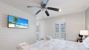 A bedroom with a bed, a ceiling fan, a TV on the wall, a window with blinds, and a nightstand with a lamp.