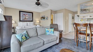 A living room with a grey sofa, a dining table with chairs, a ceiling fan, and a wall with framed pictures.