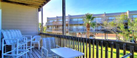 A wooden deck with white outdoor furniture, including chairs and a table, overlooking a building with multiple balconies and a clear sky.