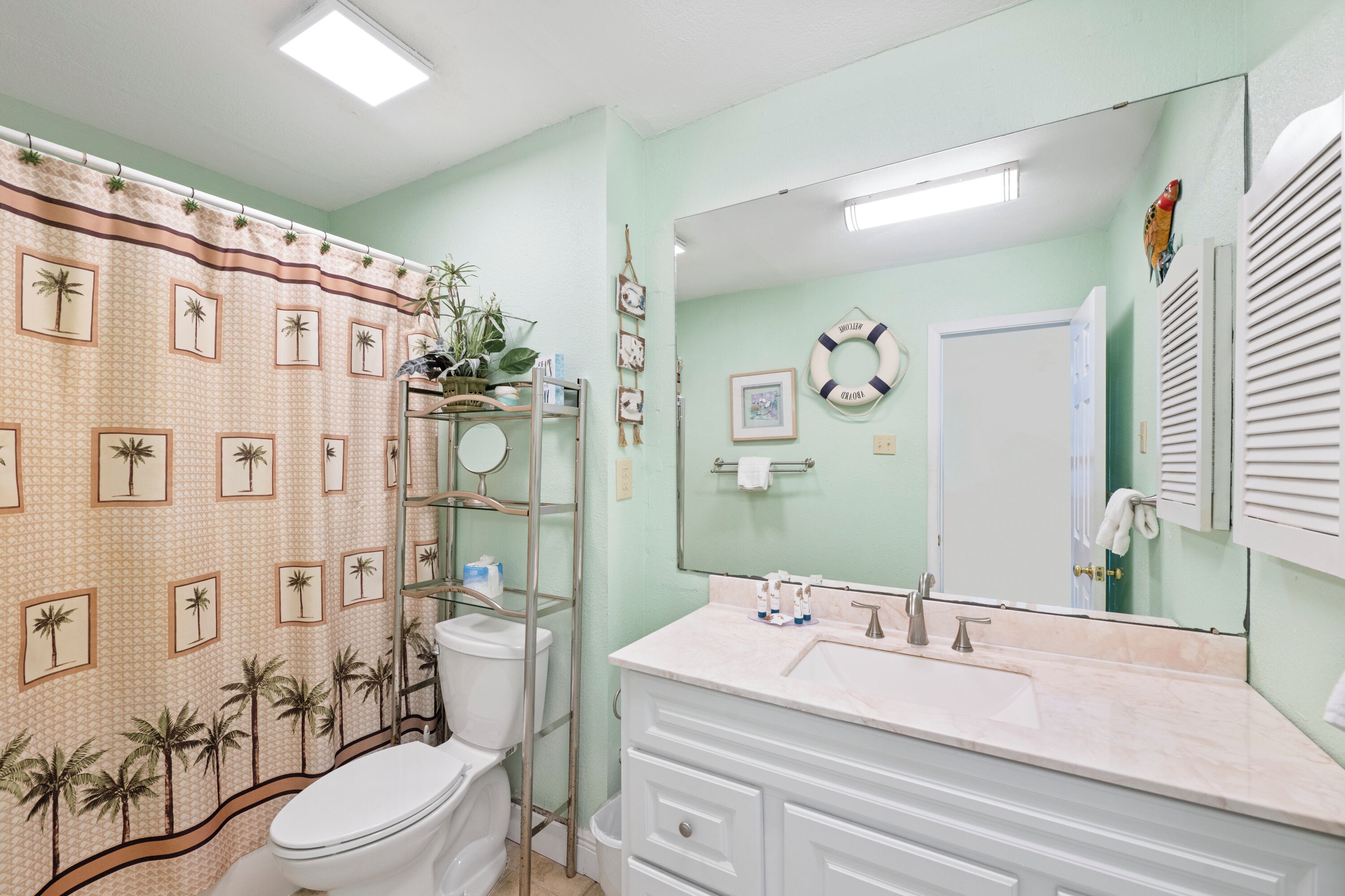 Een badkamer met toilet, wastafel en spiegel. Er is een douchegordijn met palmboommotieven.