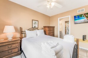 A neatly made bed with white linens and towels, a bedside lamp, a wooden nightstand, a framed picture, and a television on the wall.