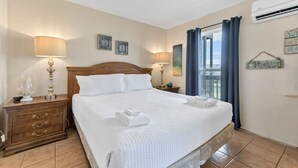 A neatly made bed with white linens and a folded towel, a wooden nightstand with a lamp, and a view of the outdoors through a window with blue curtains. - Spacious studio condo with the best amenities in Port Aransas!  Community pool, (Port Aransas)