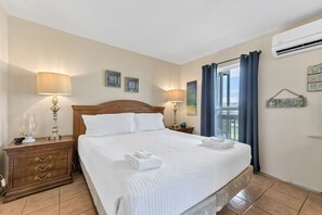 A neatly made bed with white linens and a folded towel, a wooden nightstand with a lamp, and a view of the outdoors through a window with blue curtains.