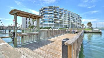 Un muelle de madera que conduce a un moderno hotel de varios pisos con balcones, un muelle y un cielo azul despejado.