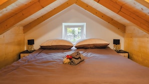 Une chambre avec un plafond en pente, une fenêtre, deux tables de chevet avec des lampes, un lit avec une couverture et une serviette ornée d’une fleur.