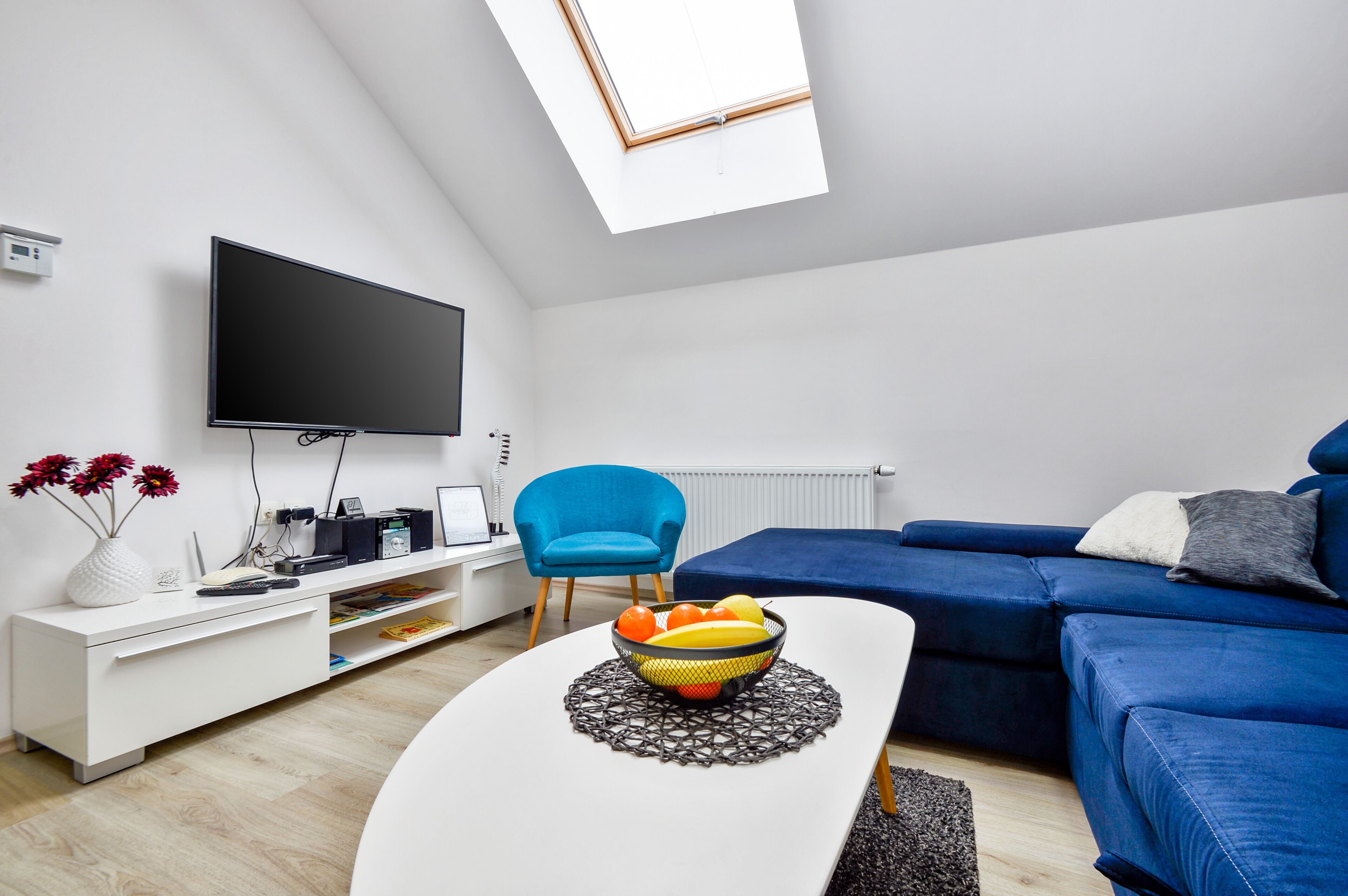 Una sala moderna con una televisión de pantalla plana, un sillón azul, un sofá azul y una mesa de comedor con un frutero.