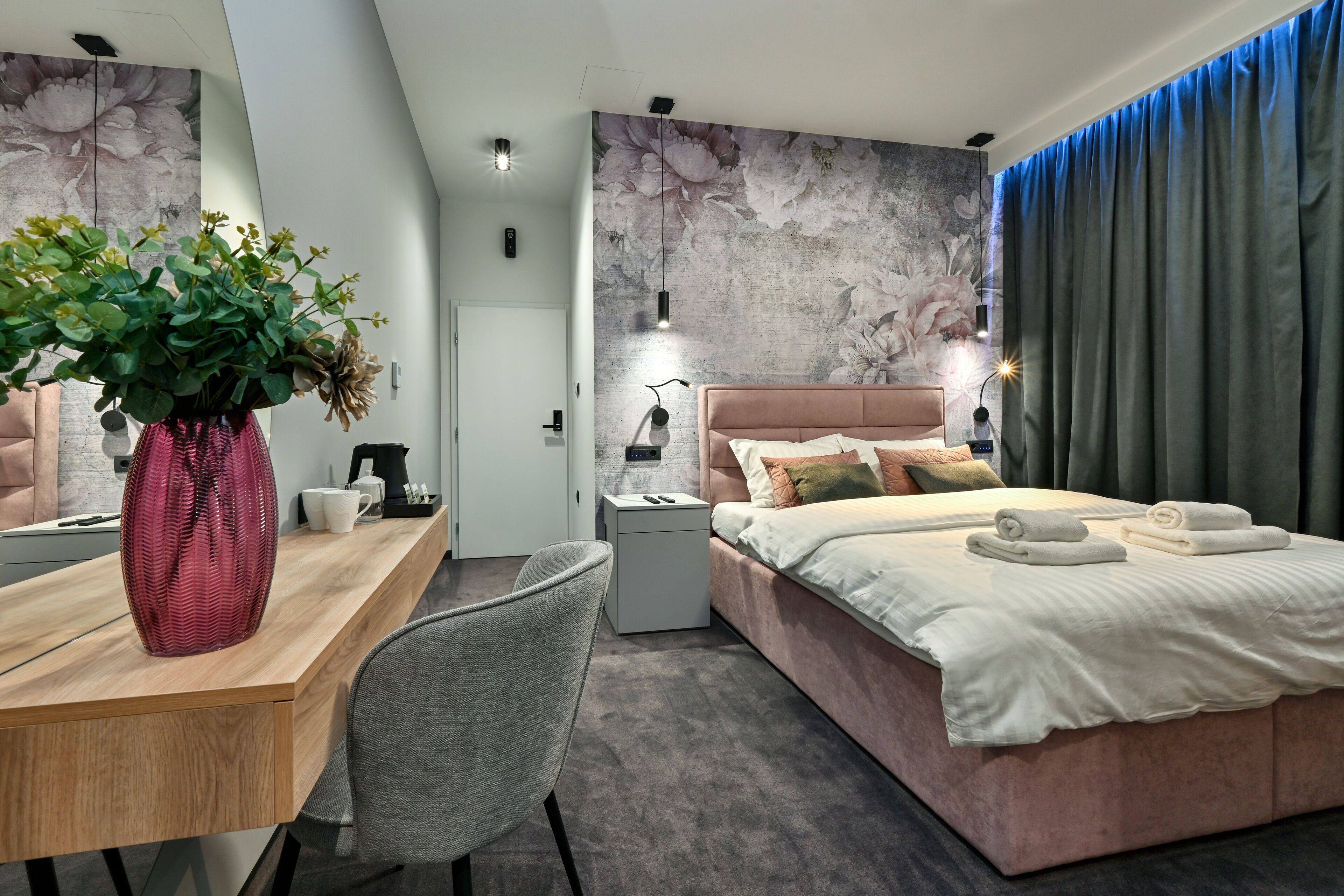 Una habitación de hotel moderna con un escritorio de madera, una silla gris, una cama con cabecera rosa y un gran mural floral en la pared.