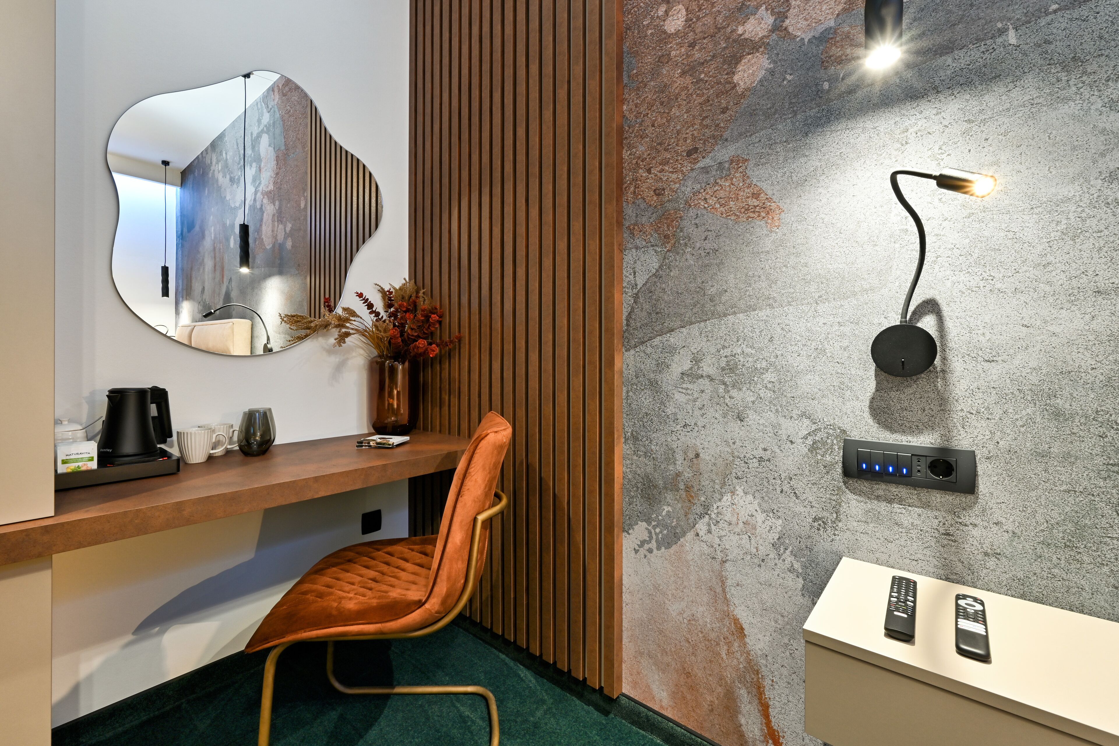 A modern hotel room with a wooden desk, a mirror, a chair, a vase with flowers, a wall-mounted lamp, and a built-in charging station.