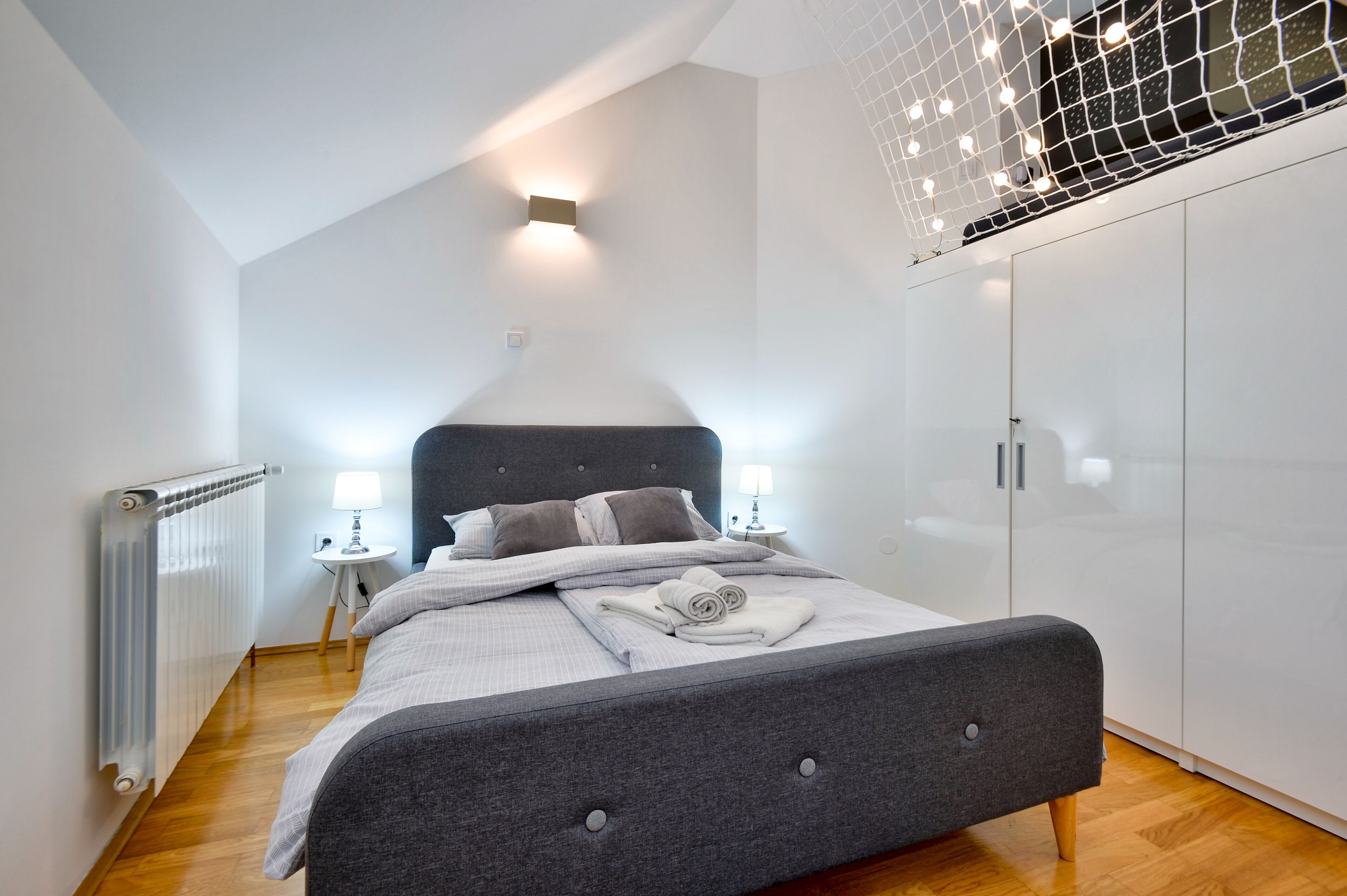 Un dormitorio moderno con una cama tapizada en gris, paredes blancas, suelo de madera, un radiador blanco, una mesita de noche con una lámpara y un armario con puertas espejadas.