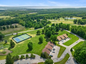 An aerial view of a residential area with a tennis court, a building with a parking lot, and a lake in the background.