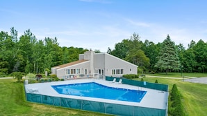 Een modern huis met een groot rechthoekig zwembad, omringd door een groene hekken.