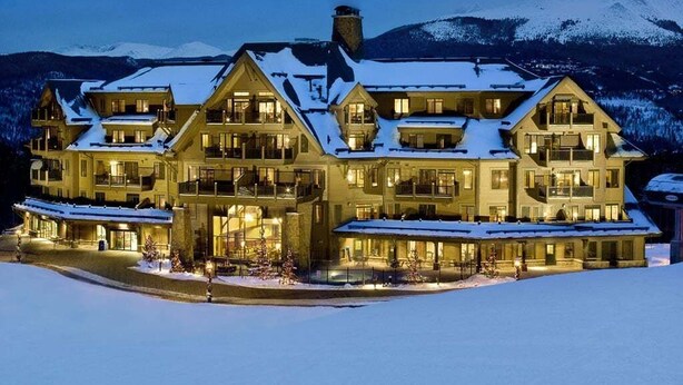 Un hotel grande de varios pisos con tejado cubierto de nieve y múltiples balcones, rodeado de un paisaje nevado y montañas.