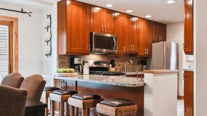 Una cocina moderna con armarios de madera, un microondas de acero inoxidable y una encimera de granito. Hay tres taburetes de madera en la barra.