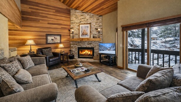 A cozy living room with a stone fireplace, wooden ceiling, and a large window overlooking a snowy landscape. - 1791 Lakeside Getaway At Deer Valley! Outdoor Heated Pool & Hot Tubs! 2 Bedroom Condo by RedAwning (Park City)