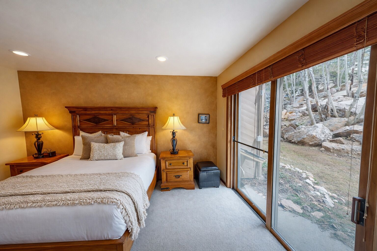 A bedroom with a large bed, two bedside tables with lamps, a small nightstand, and a view of a rocky outdoor area through large sliding glass doors.