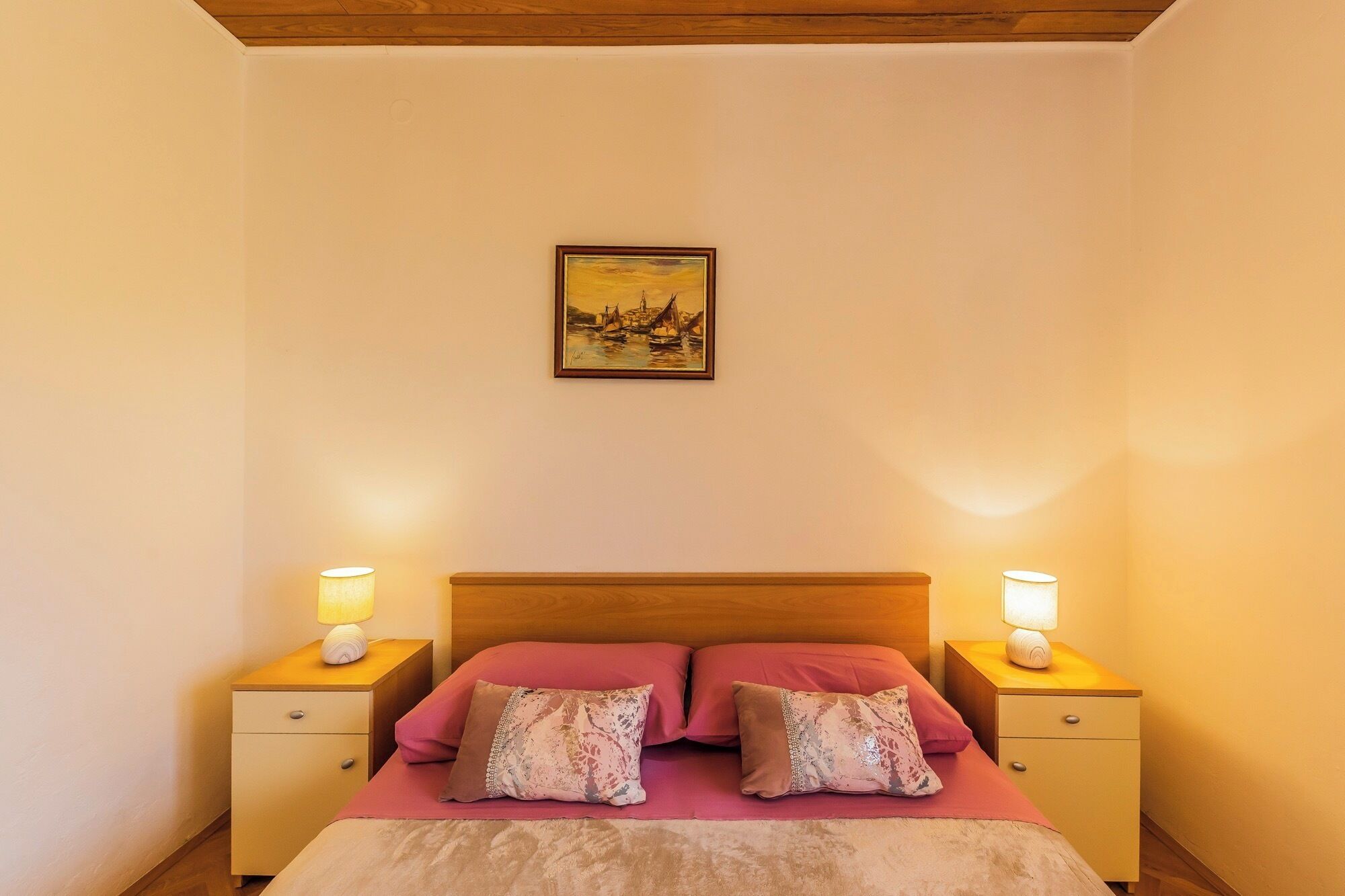 Quarto com cabeceira de madeira, duas mesas de cabeceira com luminárias, cama com roupa de cama rosa e almofadas decorativas, e um quadro na parede.