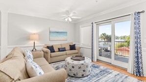 A living room with a beige sofa, a round ottoman with nautical prints, a floor lamp, and a view through a glass door to the outside.