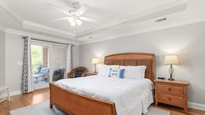 A bedroom with a wooden bed, two bedside tables with lamps, a ceiling fan, and a view through a glass door to an outdoor seating area.