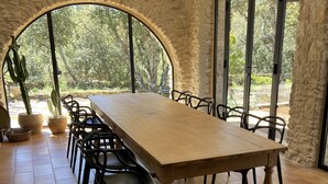 Ein langer Holztisch mit schwarzen Stühlen vor einer Steinwand, durch ein großes Rundbogenfenster mit Blick auf einen Garten.