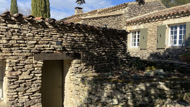 Ein Steinhaus mit Holztür und Fensterläden, umgeben von Grünpflanzen und einer Steinmauer.