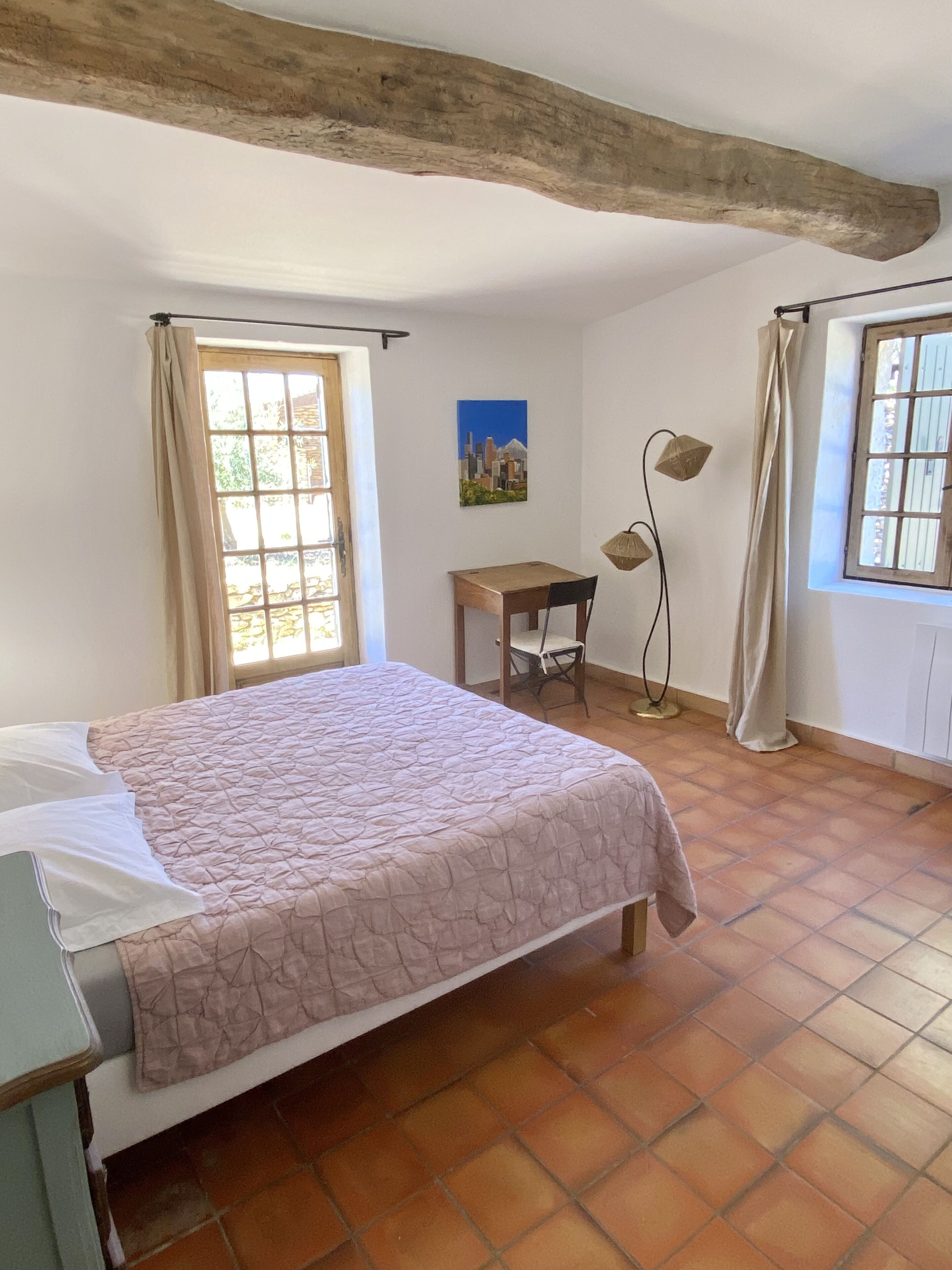 A bedroom with a bed, a desk, and a chair. There is a window with curtains, a painting on the wall, and a lamp.